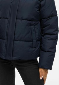 Tumesinise puffer-tagi, millel on kastiline disain, lukuga sulgemine, küljetaskud ja soonikuga mansetid, valmistatud läikivast ja kergest kangast.