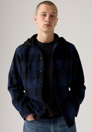 Jeune homme aux cheveux courts portant un sweat à capuche noir sous une chemise à carreaux bleue et noire boutonnée et un jean bleu, debout les mains dans les poches.