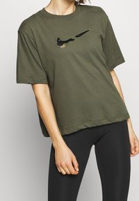 Camiseta de algodón verde oliva con un logo de swoosh de Nike en negro y camuflaje, corte holgado, mangas cortas y cuello redondo.