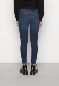 Jeans de denim escuros, skinny fit, com bolsos traseiros, combinados com botas pretas com costuras e atacadores amarelos, de pé sobre um chão cinzento.