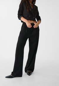 Vrouw in een zwarte cropped blazer en hoge taille brede broek, die de voorknoop aanpast, met puntige zwarte schoenen met metalen oogjes.