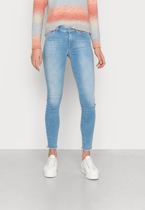 Femme portant un jean skinny bleu clair avec des ourlets effilochés, un pull rayé multicolore et des baskets blanches à plateforme, debout sur un sol gris.