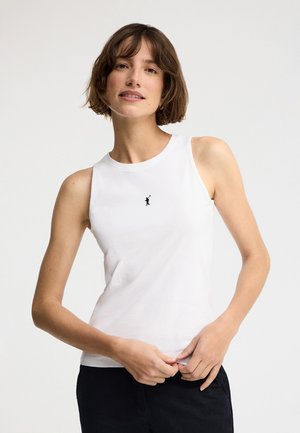 Femme aux cheveux courts et bruns portant un haut blanc sans manches avec un petit logo de golfeur noir, ajustant l'ourlet, sur un fond clair uni.