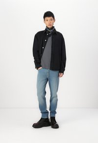 Camisa negra de botones sobre una camiseta gris, combinada con jeans azul claro y botas negras. Se lleva un pañuelo de patrón blanco y negro alrededor del cuello.
