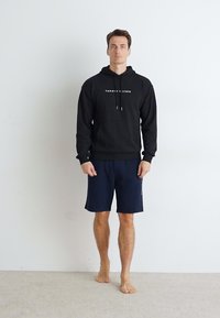 Svart hettegenser med hvit logo, kombinert med marineblå shorts. Modellen står barføtt på lys teppe foran en ensfarget veggbakgrunn.
