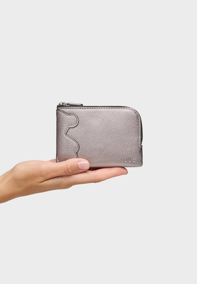 Dłoń trzymająca mały metaliczny srebrny portfel zapinany na zamek z teksturowaną powierzchnią i subtelnym wytłoczonym logo marki.