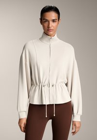 Lysbeige cropped jakke med høj krave, fuld lynlås, snoreliv og sænkede skuldre. Glat stof, blød tekstur, minimalistisk design.