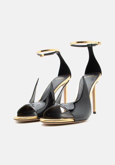 Μαύρα και χρυσά ψηλοτάκουνα πέδιλα με ανοιχτά δάχτυλα και λουράκια στον αστράγαλο, με γυαλιστερό φινίρισμα, τοποθετημένα δίπλα-δίπλα σε λευκό φόντο.