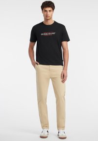 Schwarzes T-Shirt mit rundem Ausschnitt und rotem sowie weißem "GUESS JEANS"-Logo, kombiniert mit beigen gerade geschnittenen Hosen und weißen Sneakers mit schwarzen Akzenten.