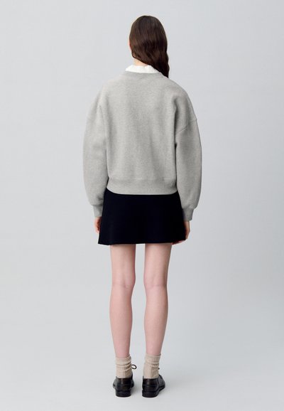 Felpa grigia con spalle scese, polsini e orlo a coste, abbinata a una gonna nera. Indossata con calzini beige e scarpe nere.