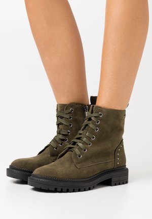 Oliivinvihreät mokkanilkkurit, joissa on nauhoitettu etuosa, sivuvetoketju, kulutusta kestävä musta pohja ja hopeanväriset niittikoristeet ulkosyrjällä.