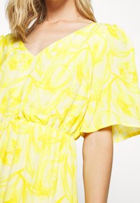 Robe florale jaune avec un décolleté en V et des manches courtes et larges. Le tissu présente un motif texturé, peint à la main, de fleurs et de lignes tourbillonnantes.