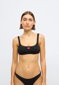 Črni bikini zgornji del z rebrasto teksturo, kvadratnim izrezom in širokimi naramnicami, z majhno rdečo logotipno značko na sredini. Gladek otip, oprijet dizajn.