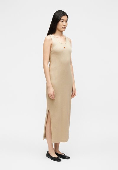 Γυναίκα που στέκεται με μπεζ midi φόρεμα χωρίς μανίκια, με πλαϊνές σκισίματα, μικρή κεντημένη τίγρη στο στήθος και μαύρα επίπεδα παπούτσια, πάνω σε λευκό φόντο.