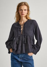 Blusa grigia ricamata con scollatura lacciata, maniche lunghe, frange decorative e orlo scalloppato. Realizzata in tessuto leggero.