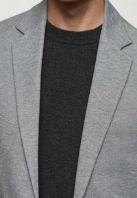 Blazer grigio chiaro in tessuto testurizzato con rever a punta, indossato sopra un maglione a collo rotondo grigio scuro. Dettaglio a colletto visibile all' neckline.