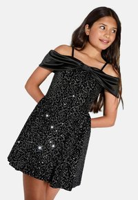 Melna kleita ar plecu augšdaļu un satīna loku, rotāta ar mirdzošām zeltīšiem. Pieguļošs augšdaļa, paplatināta svārku daļa un regulējamas siksnas.