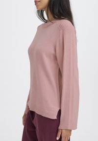Jasnoróżowy sweter z miękkiego materiału, z prążkowanymi detalami na ramionach i rękawach, prostym dołem oraz luźnym krojem.