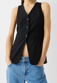 Svart väst med smalrandigt mönster och V-ringning, med tre knappar, sidofickor och en skräddarsydd passform, bärs över ljusblå högmidjade jeans.