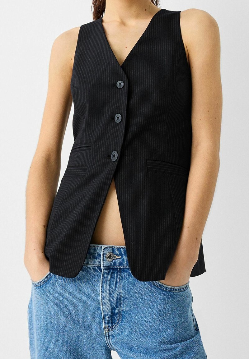 Svart väst med smalrandigt mönster och V-ringning, med tre knappar, sidofickor och en skräddarsydd passform, bärs över ljusblå högmidjade jeans.