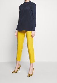 Granatowa bluzka z długim rękawem i kołnierzykiem mandarynkowym, w połączeniu z jasnymi, żółtymi dopasowanymi spodniami oraz czarno-żółtymi butami na obcasie w paski. Prosty, nowoczesny design.