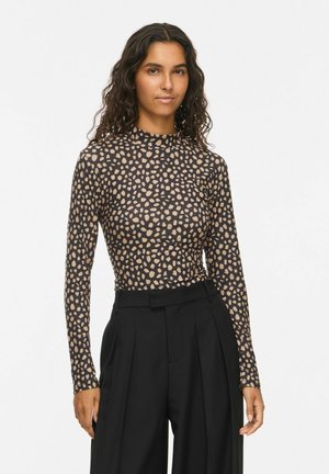 Femme aux longs cheveux bouclés porte un haut à manches longues noir et beige à pois, associé à un pantalon noir plissé taille haute.