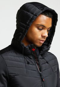 Giubbotto imbottito nero con cappuccio trapuntato, tasche laterali e dettagli rossi. Presenta una chiusura con zip e una finitura in tessuto liscio.