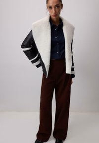 Sieviete, kas valkā melno un baltā vilnas jaku, tumšo džinsu kreklu ar pogām, brūnas platu kāju bikses un melnas kurpes uz vienkārša fona.