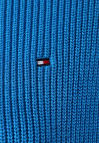 Dzianina w kolorze niebieskim z pionowym prążkowaniem. Obecny jest mały haftowany logo z czarnymi, białymi i czerwonymi paskami. Miękka tekstura.