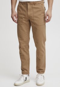 Tan katoen cargo pants met een rechte pasvorm, voorzien van twee voorzakken en een knopenvluiting. Gecombineerd met witte sneakers met zwarte accenten.