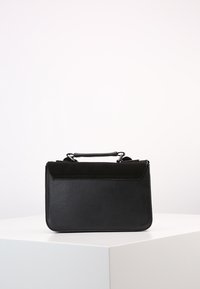 Borsa rettangolare nera con manico superiore, posata su un piedistallo bianco contro uno sfondo chiaro e semplice.