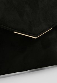 Pochette enveloppe en daim noir avec un détail métallique en forme de V doré sur le rabat, présentée sur une surface gris clair.