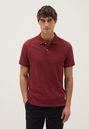 Jonge man met kort bruin haar, gekleed in een aangesloten donkerrood poloshirt met korte mouwen en een zwarte broek, staand met handen in de zakken.