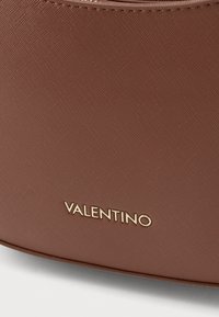 Bolsa de couro texturizado castanho com uma forma curvada, apresenta um logo dourado "Valentino" na parte inferior. Superfície lisa com um leve brilho.
