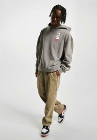 Felpa grigia con tasca frontale, grafica del logo, abbinata a pantaloni di corda beige larghi e sneakers nere con dettagli rossi.