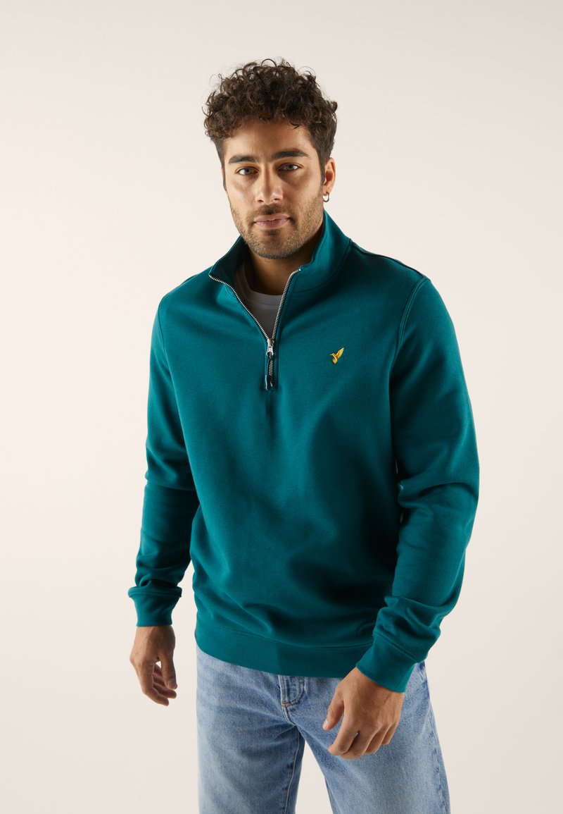 Teal kvartszipsweatshirt med mjuk textur, utrustad med en gul logotyp på bröstet och muddade ärmslut, kombinerad med ljusblå jeans.