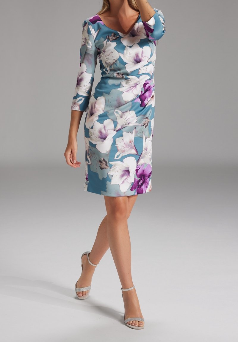 Femme portant une robe bleue mi-longue avec de grandes fleurs blanches et violettes, debout, une jambe croisée devant.