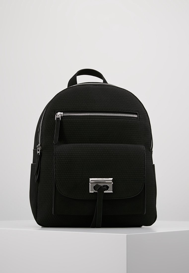 Sac à dos noir texturé avec poignée supérieure, poche avant zippée, poche à rabat avec fermoir argenté et tirettes en cuir, sur un piédestal blanc.