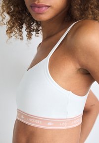Bralette blanche en tissu doux, avec des fines bretelles et un élastique rose clair portant le logo 'LACOSTE'. Texture lisse, design minimaliste.