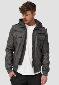 Chaqueta de cuero gris con capucha, con bolsillos con cremallera, puños acanalados y un acabado texturizado. Llevada sobre una camisa blanca.