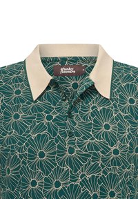 Polo verde con flores y cuello beige claro. Presenta un patrón floral blanco y tres botones en el escote. Tejido suave y texturado.