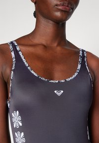Schwarzer Badeanzug mit weißen Blumenakzenten und gemusterten Trägern. Verfügt über einen runden Ausschnitt und ein kleines Logo auf der Brust. Sanfte Textur.