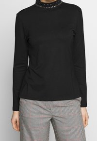 Svart långärmad topp med en figurnära silhuett, med en strukturerad krage prydd med cirkulära dekorationer. Mjukt material, enkel design.