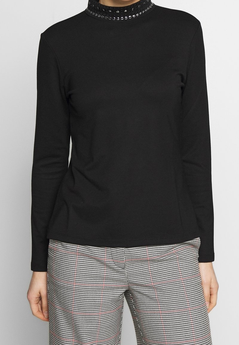 Svart långärmad topp med en figurnära silhuett, med en strukturerad krage prydd med cirkulära dekorationer. Mjukt material, enkel design.