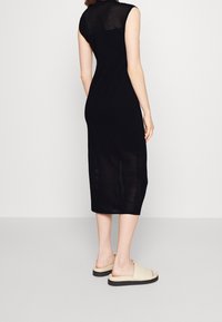 Robe noire sans manches présentant un motif côtelé sur la moitié supérieure, se transformant en une section inférieure unie. Le mannequin porte des sandales de couleur claire.