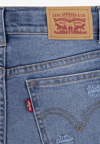 Vaqueros de mezclilla azul claro con una etiqueta de cuero marrón, costuras visibles y una pequeña etiqueta roja en el bolsillo trasero. Presentan un aspecto desgastado.