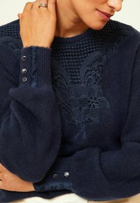 Pull en maille bleu marine avec broderie florale sur la poitrine, tissu texturé et boutons décoratifs sur les manches.