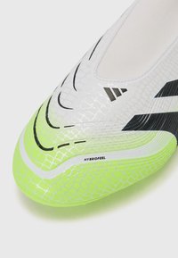 White soccer cleat with textured upper, featuring black stripes and a bright green sole. "HYBRIDFEEL" label visible near the toe.