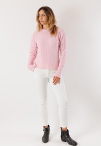 Donna dai capelli ondulati, che indossa un maglione rosa a trecce, pantaloni bianchi e stivaletti neri alla caviglia, in piedi con le mani in tasca e lo sguardo rivolto di lato.