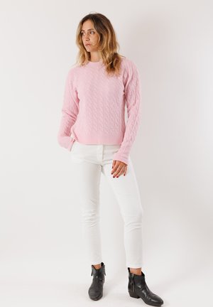 Donna dai capelli ondulati, che indossa un maglione rosa a trecce, pantaloni bianchi e stivaletti neri alla caviglia, in piedi con le mani in tasca e lo sguardo rivolto di lato.